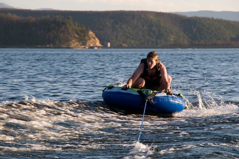 Teenage boy riding an inflatable tube being towed behind a boat on a lake in summer.