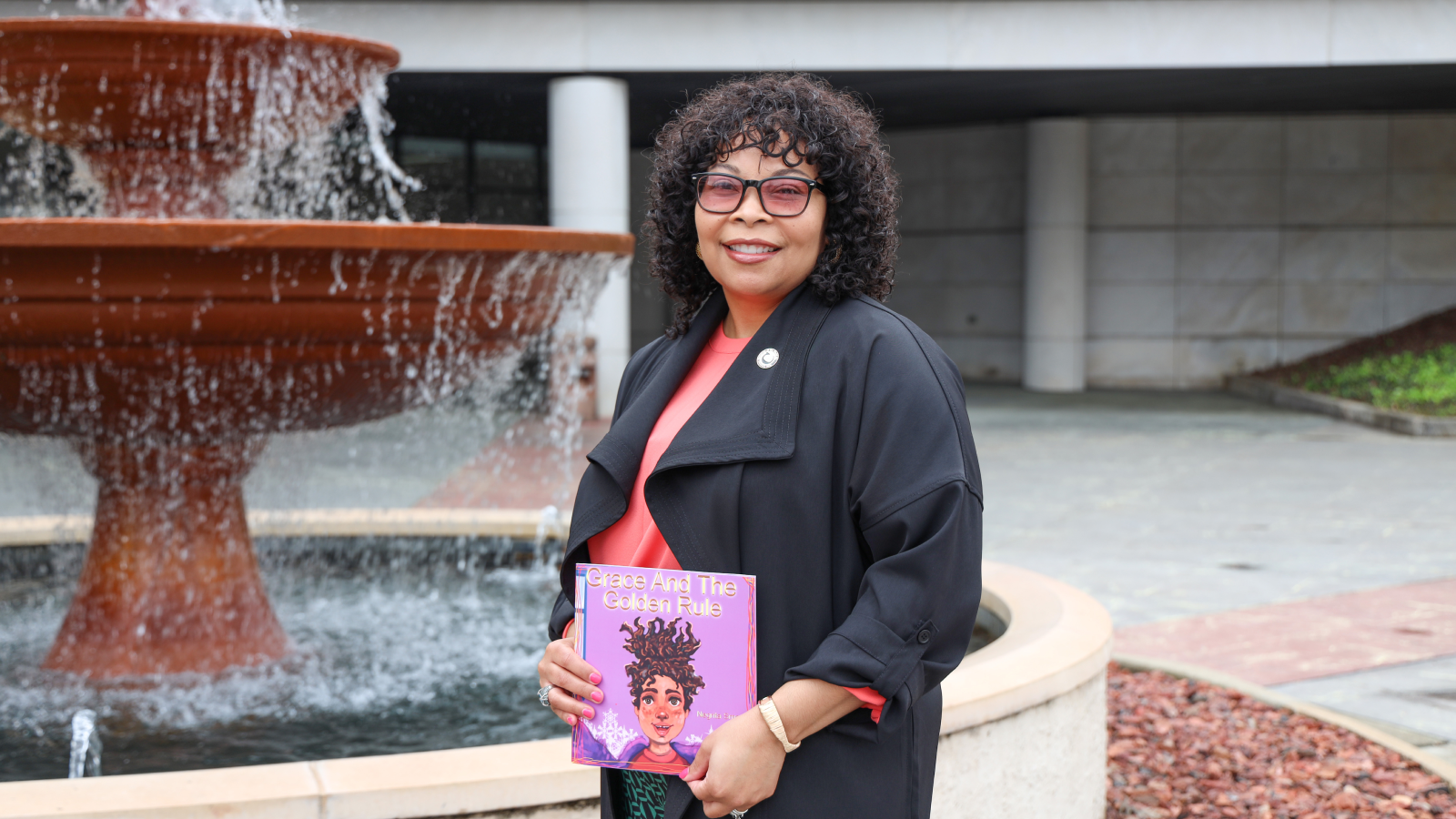 Negrita Smart holding her book, Grace and The Golden Rule, standing in front of a fountain.