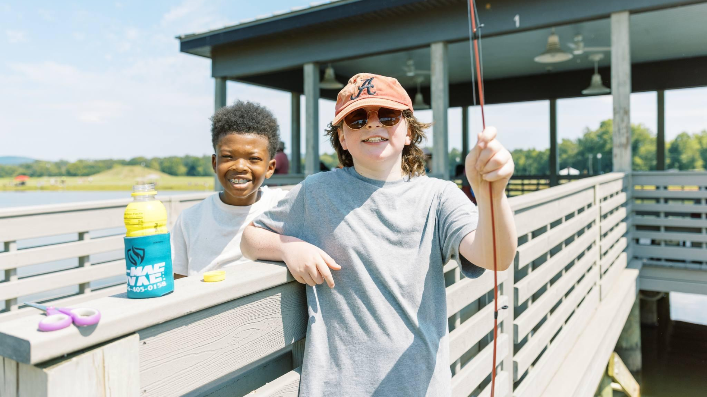 Two young boys standing on dock with fishing supplies.