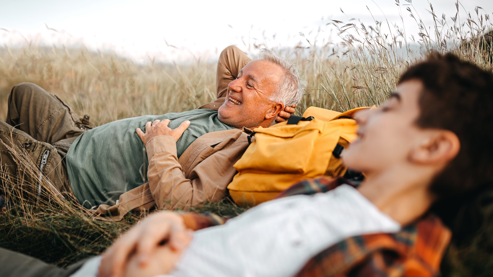 Grandfather smiling beside his grandson as they lie in tall grass and rest on their hiking backpacks.