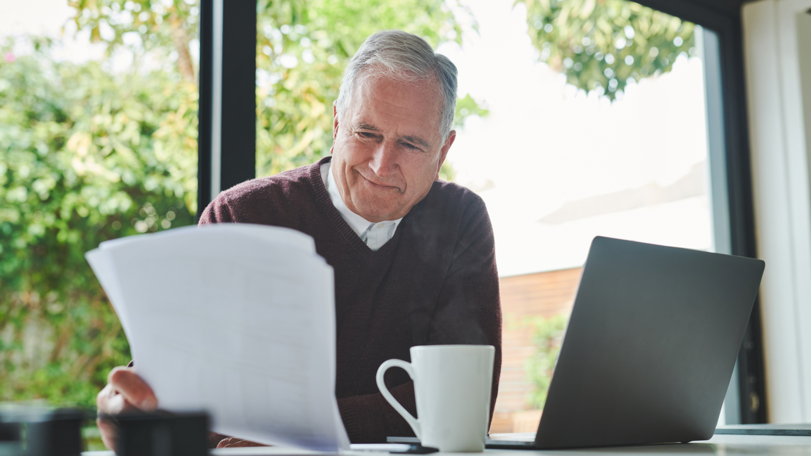 Older man in a red shirt reviewing homeowners insurance policy documents at his computer with a cup of coffee, smiling as he understands his coverage options
