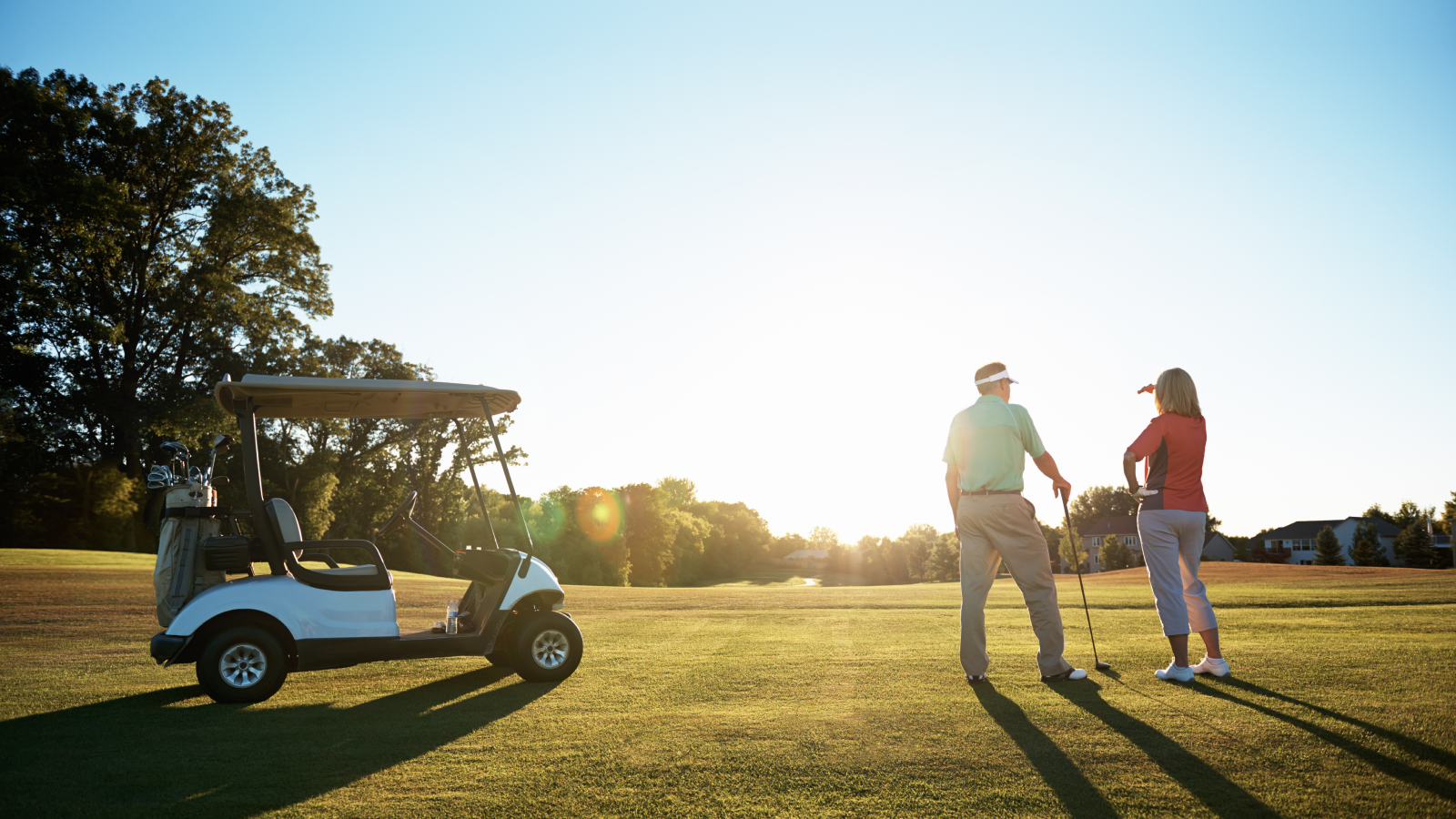 A retired couple stands beside their golf cart on a fairway at sunset.