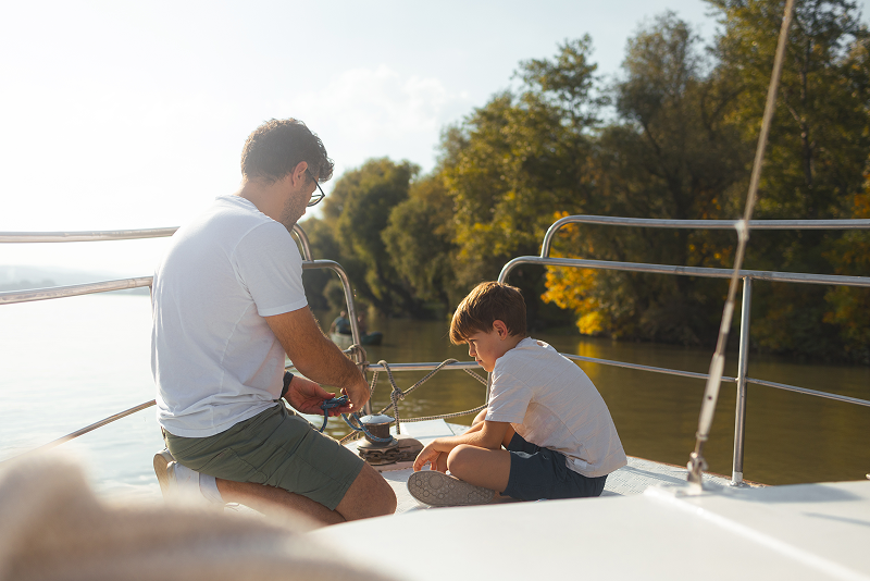 A young boy sitting on the bow of a boat learns to tie a boater’s knot from his father.