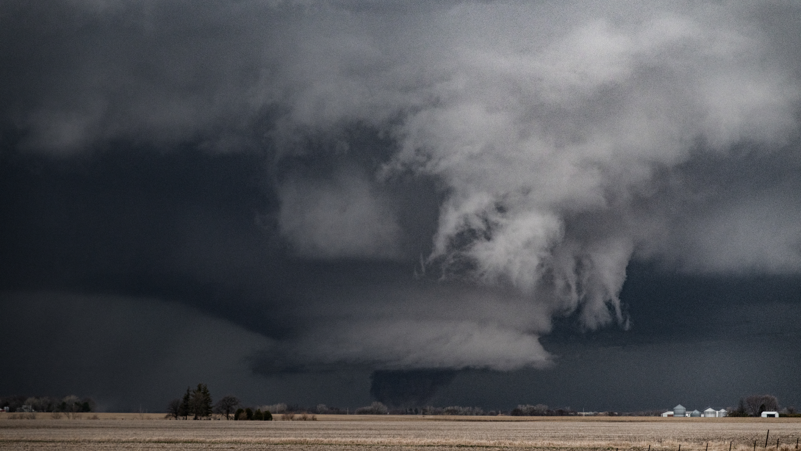 Wide tornado funnel traveling across farmland in the distance with heavy storm clouds overhead.