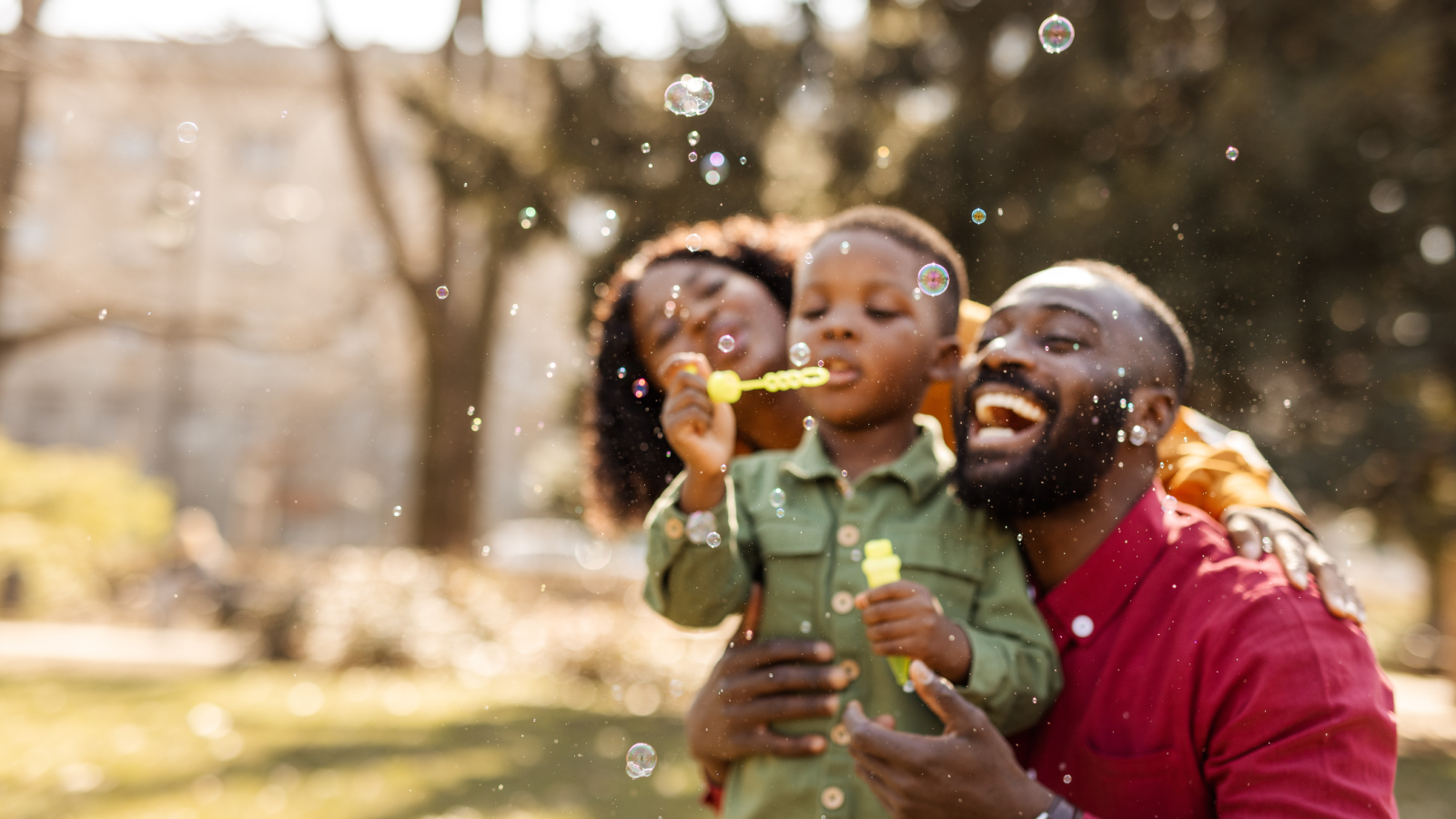 Two parents and a young boy smiling while blowing bubbles outdoors.
