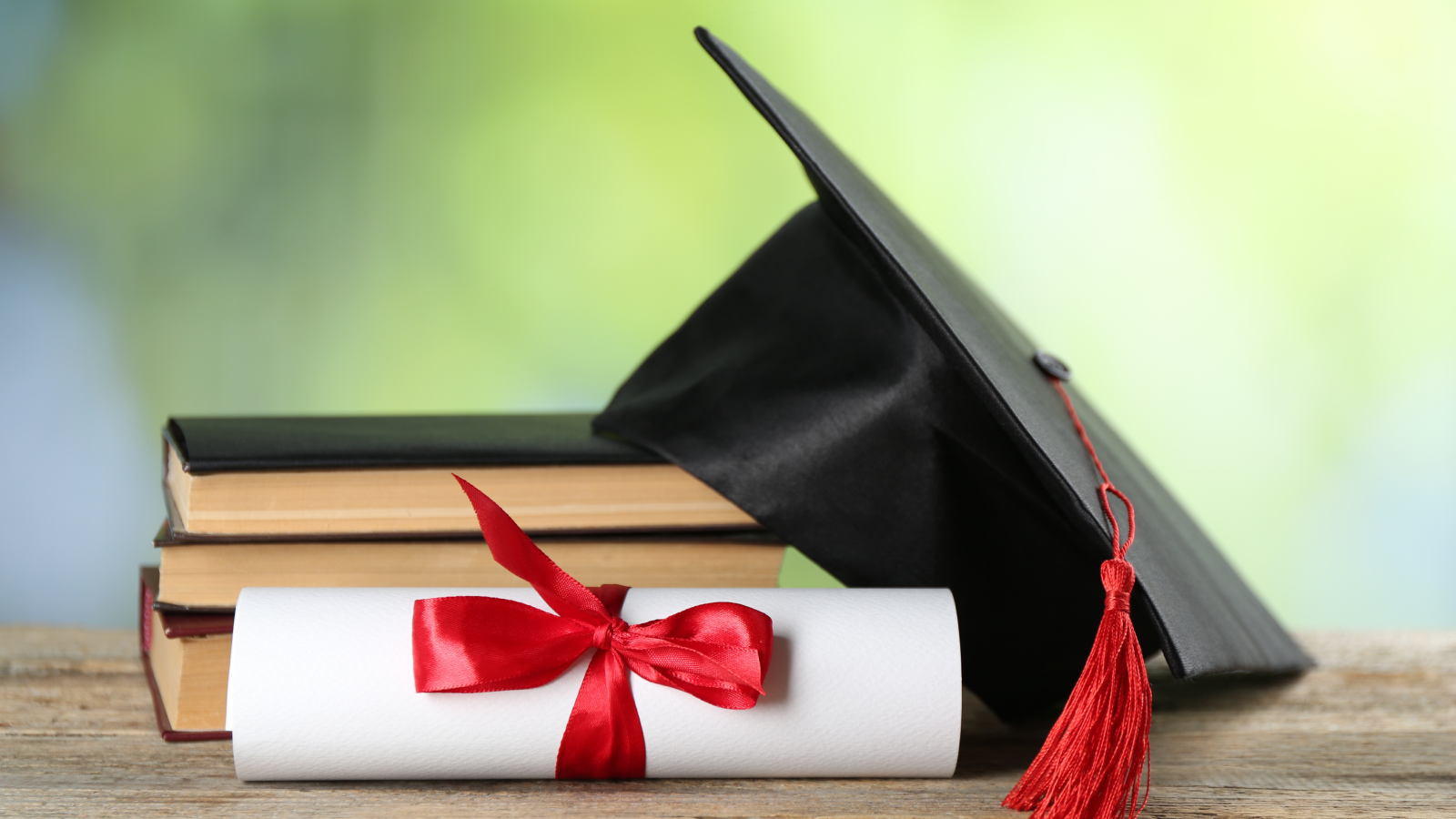 Black graduation cap resting on stacked books beside a rolled diploma tied with a red ribbon, symbolizing academic achievement.