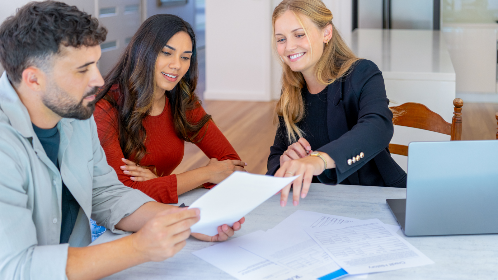 A young couple reviews home-buying paperwork with female real estate agent at kitchen table.