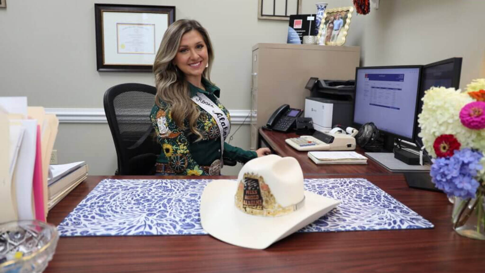 Insurance agent Emery Norred smiles at her desk, wearing a pageant sash with a cowboy hat beside her.