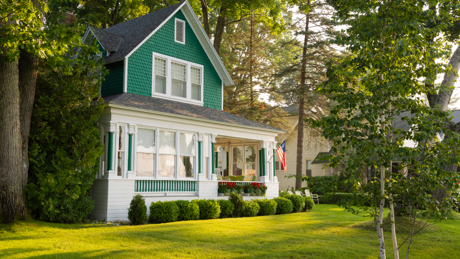 Green two-story house with a covered front porch, an American flag, mature shade trees, and a well-maintained lawn in a quiet residential neighborhood.