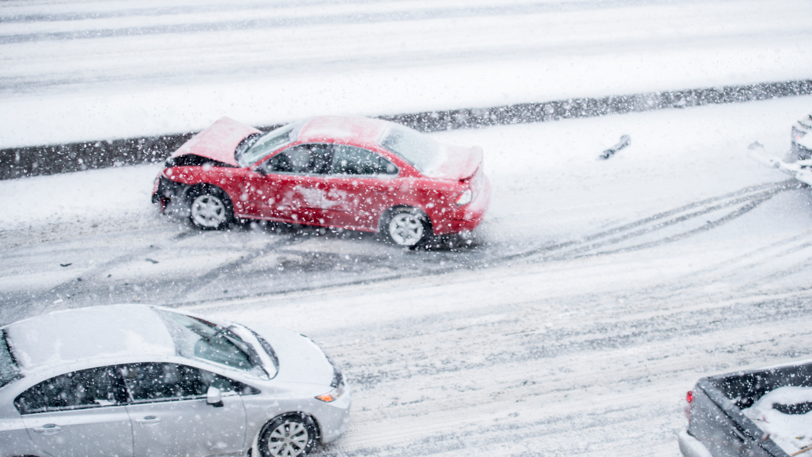 A red car with front-end damage sits stranded on an icy road after a multi-car crash, with snow falling heavily around it.