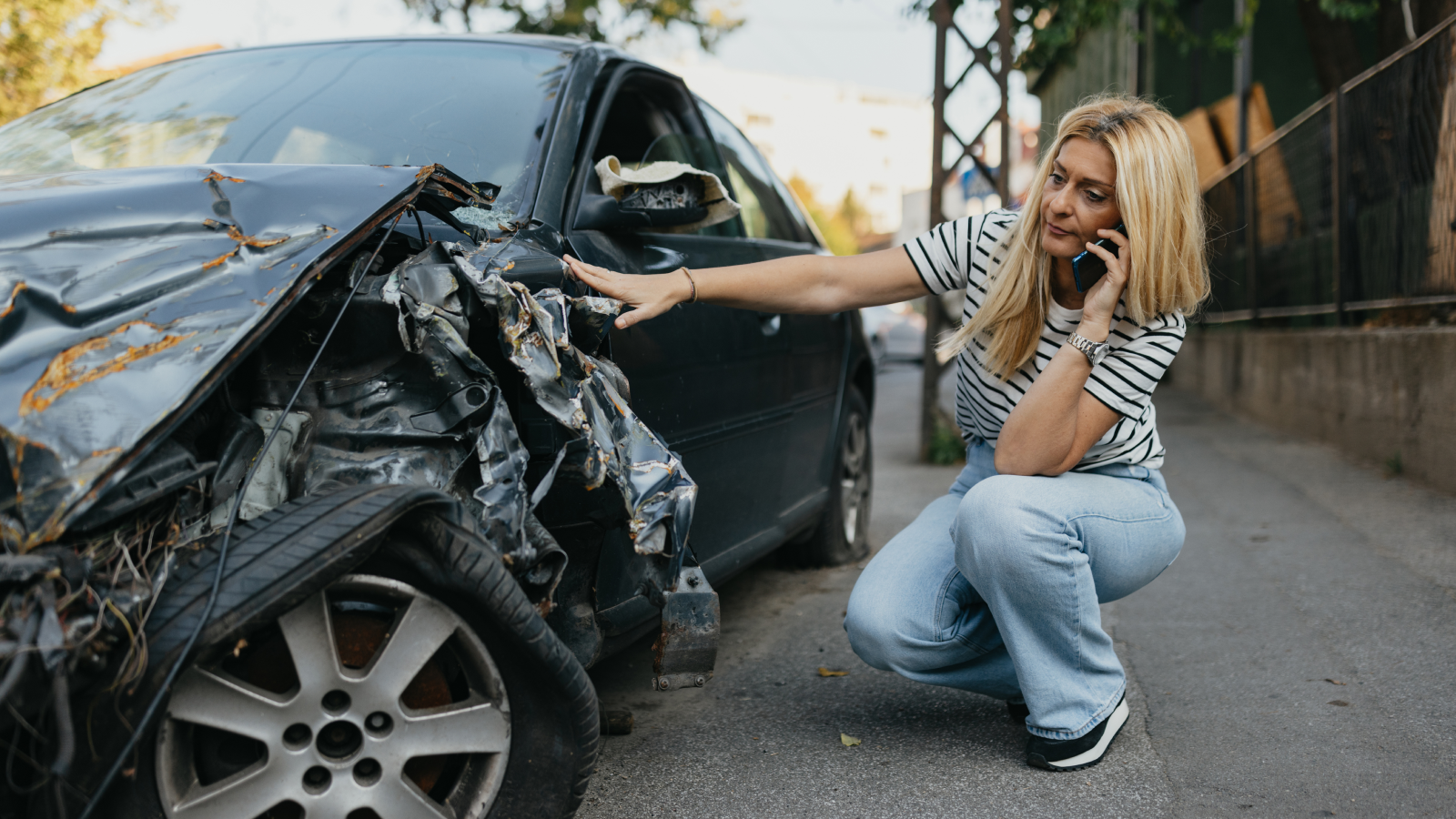 Woman kneeling beside her wrecked car after an accident, talking on her phone and assessing vehicle damage, illustrating uninsured or underinsured motorist coverage claim scenario.