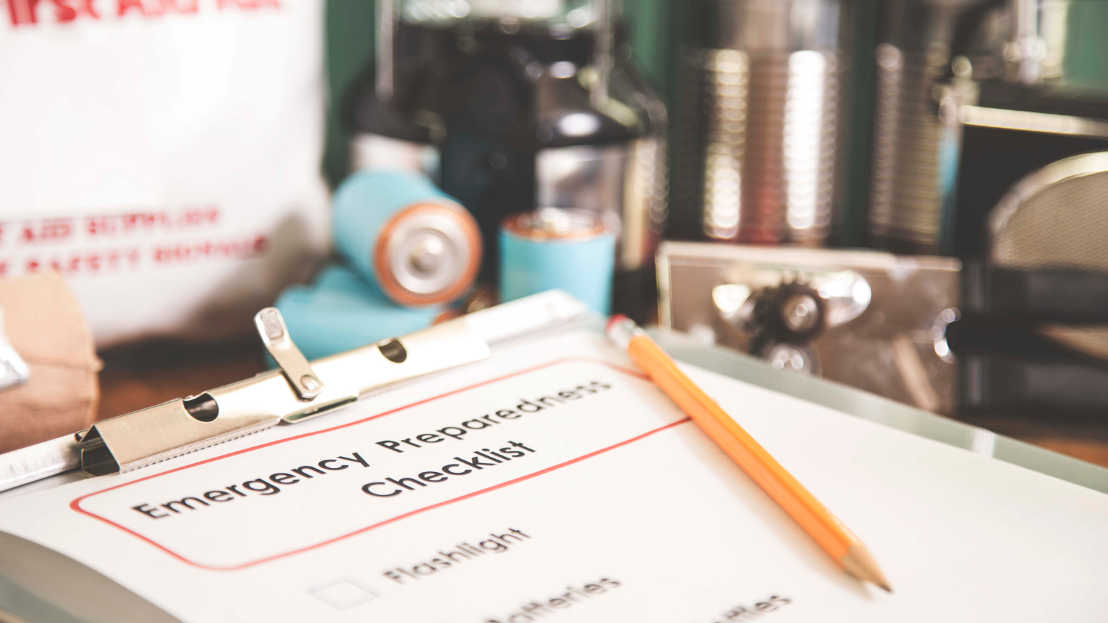 Clipboard checklist titled Emergency Preparedness with pencil, first aid kit, flashlight, and batteries in the near background